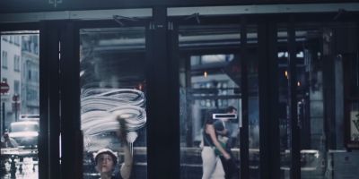 Film still from 'Rose' (2023) by Jon Beaton. A young woman is outside the cinema cleaning the windows with a sponge and soapy water. The sun is behind her, backlighting her and the soapy streaks.